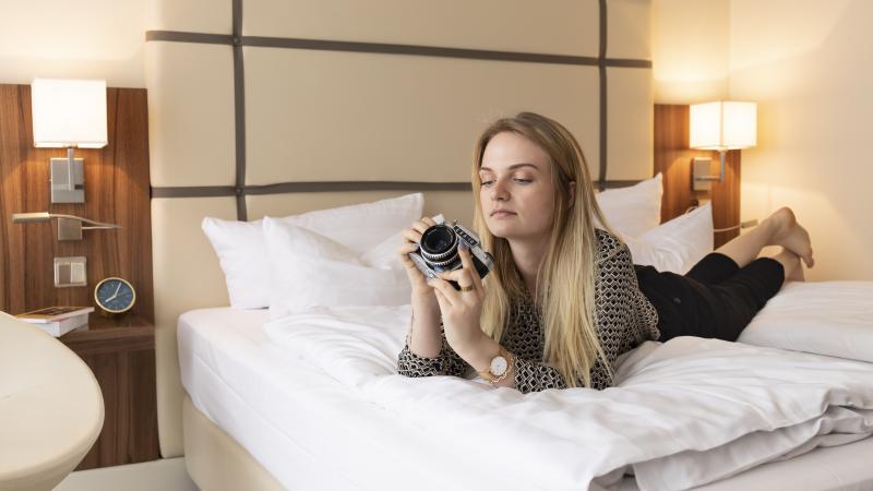 Junge Frau mit Fotokamera auf gemütlichem Bett liegend, schaut sich die Einstellungen der Kamera an. Das helle und freundliche Zimmer lädt zum entspannen und verweilen ein, auf dem Nachttisch kann man Bücher und einen Wecker sehen.