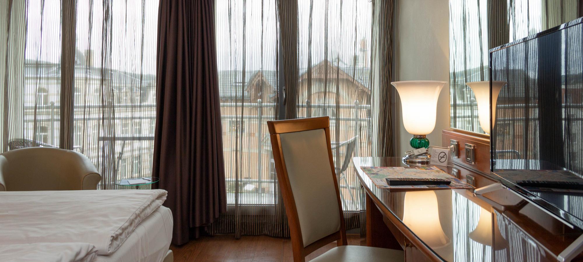 Doppelzimmer mit großzügigen, weißen Betten und Schreibtisch, mit einem cremefarbenen Sessel sowie mit Blick auf bodentiefe Fenster mit Balkon und einer Aussicht auf das Villenviertel Blasewitz in Dresden